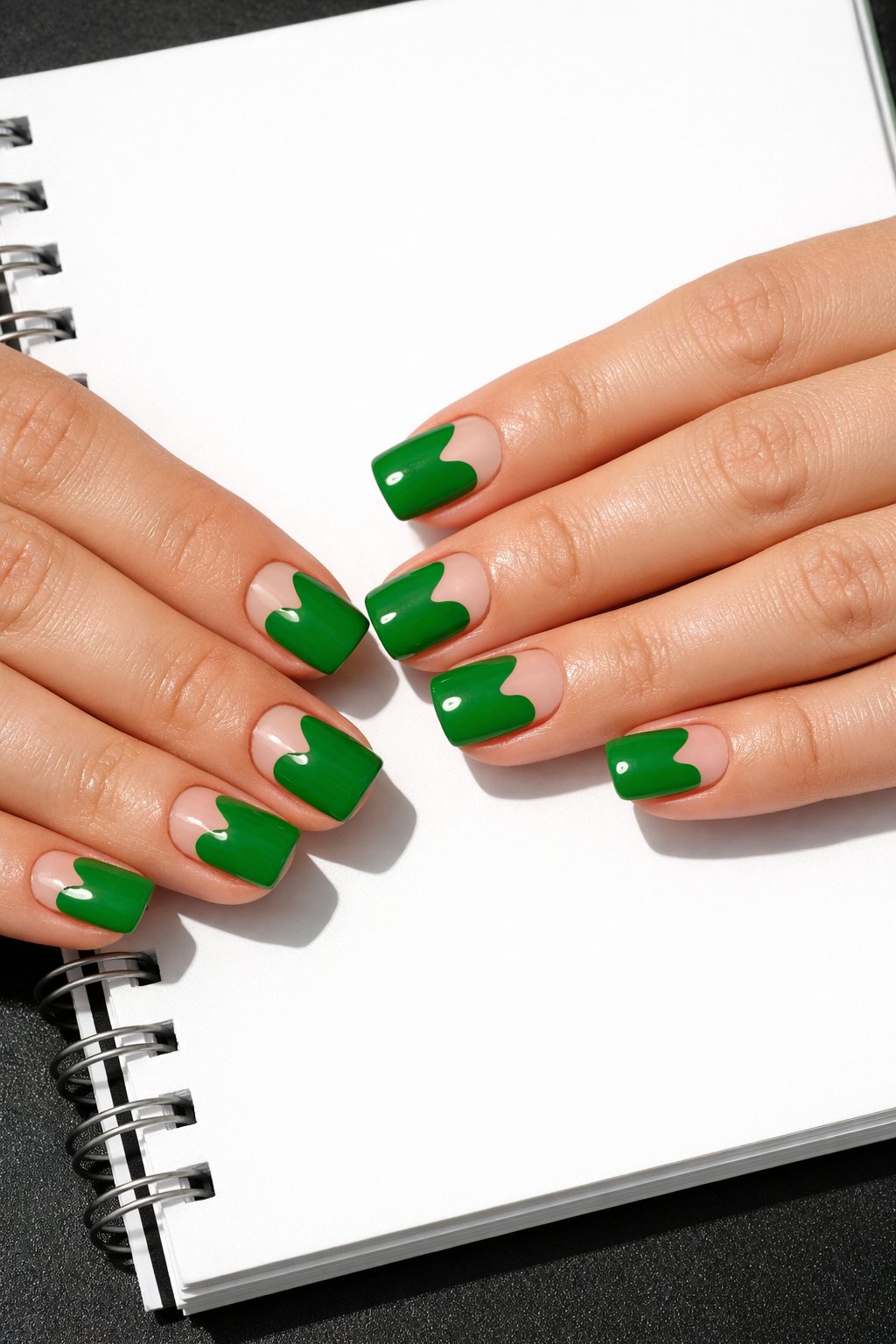 Overhead shot of hands with short, squared-off nails on a bright white notebook. The nails are a high-gloss, vibrant kelly green. Each has a crisp, heart-shaped negative space cut-out near the free edge. Direct light creates a strong, cheerful contrast. The feel is energetic and graphic.