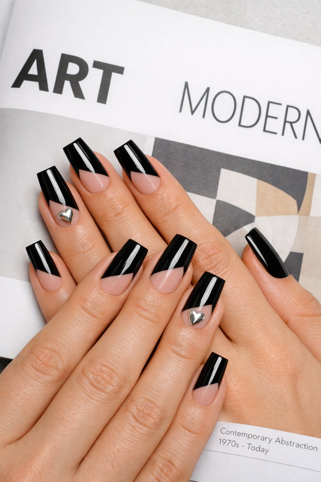 Overhead shot of hands with long, square nails resting on a modern art magazine. The design features a diagonal block of glossy black on each nail. Where the black color ends on the ring finger, a single, tiny 3D silver heart charm is attached. Clean, natural lighting keeps the look sharp. The aesthetic is editorial and sleek.
