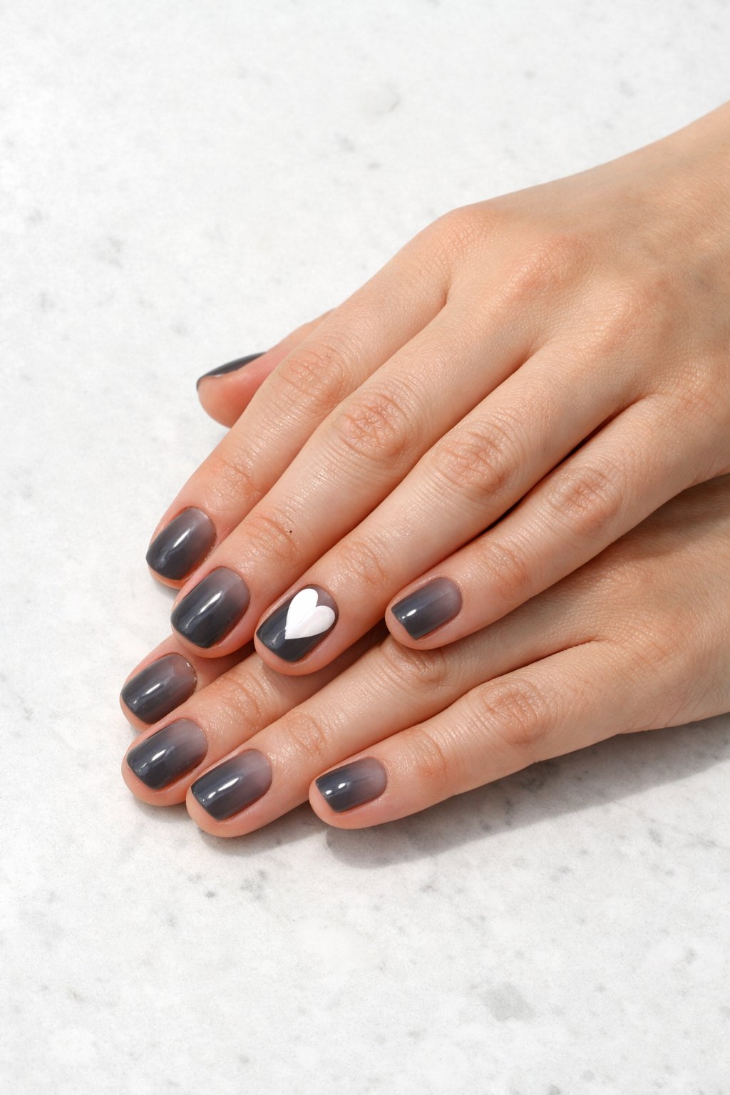 Overhead shot of hands with very short, natural nails resting on a clean, white stone countertop. The nails have a single, sheer wash of black, creating a soft, translucent grey look. On the ring finger, a bold, painterly white heart is applied with full opacity. Bright, natural light keeps the look fresh and stark. The aesthetic is minimalist and artistic.