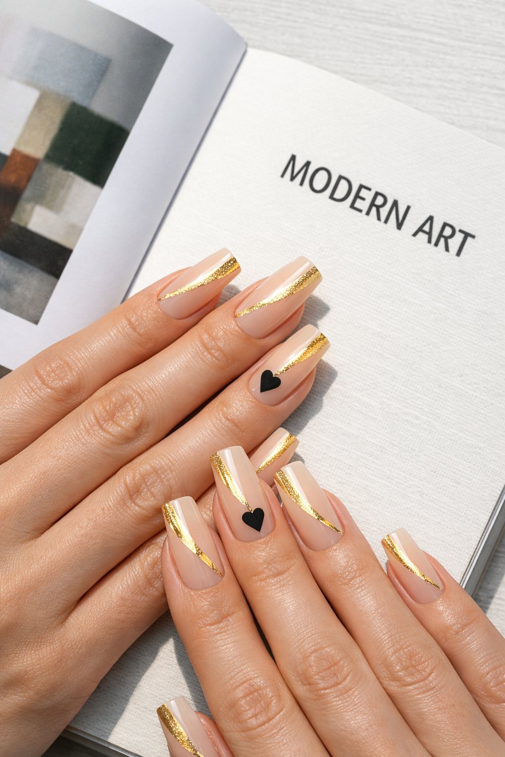 Overhead view of hands with long, square nails on a contemporary art book. Each nail has a sheer nude base with a sharp diagonal stripe of gold foil. On the ring finger, a single, tiny matte black heart sits at the apex where the gold meets the nude. Clean, natural lighting keeps the look sharp. The aesthetic is editorial and sleek.