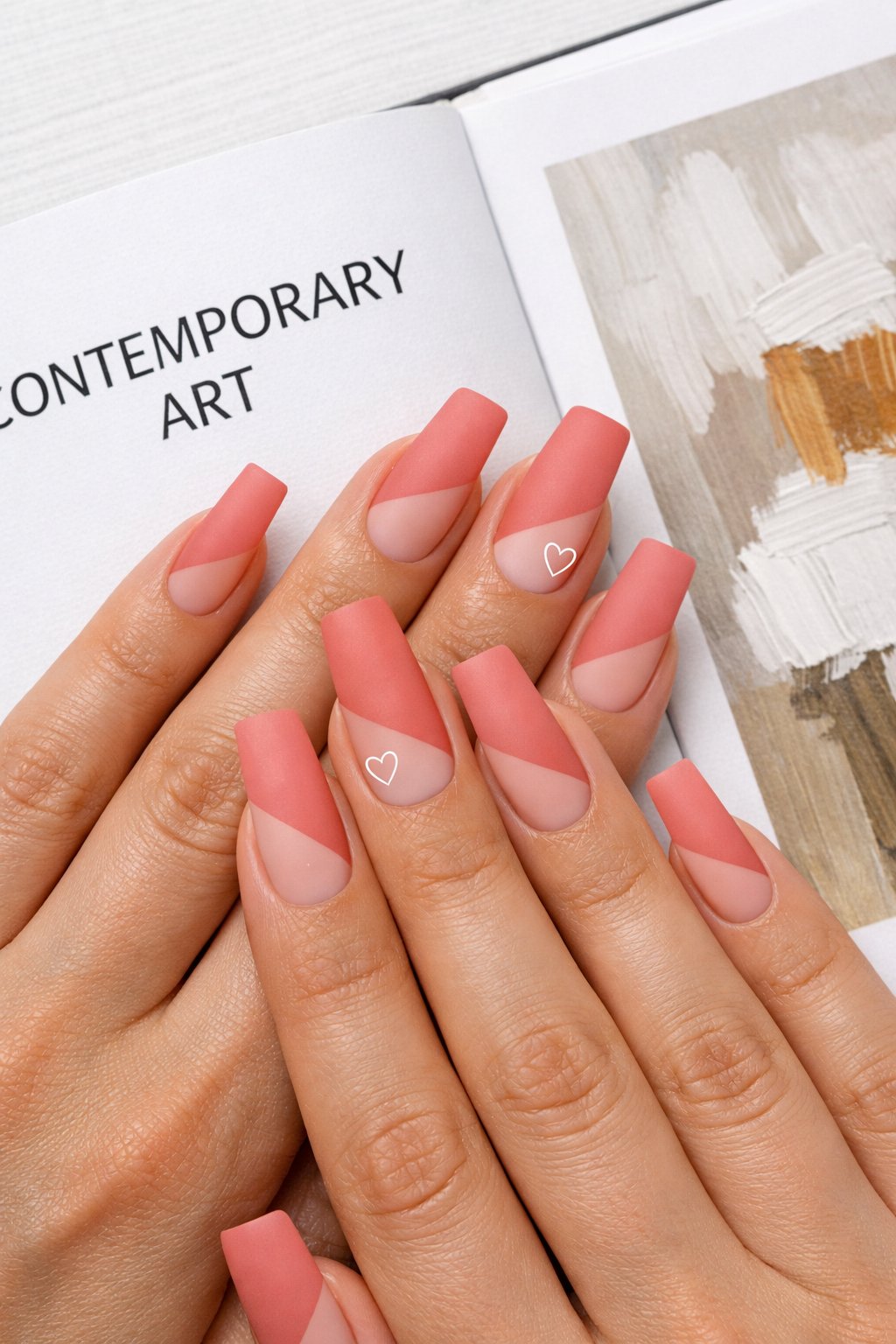 Overhead view of hands with long, square nails on a contemporary art book. The design features a diagonal block of matte salmon pink on each nail. On the ring finger, a single, fine-lined white heart sits at the border where the color ends. Clean, natural lighting keeps the look sharp. The aesthetic is editorial and sleek.