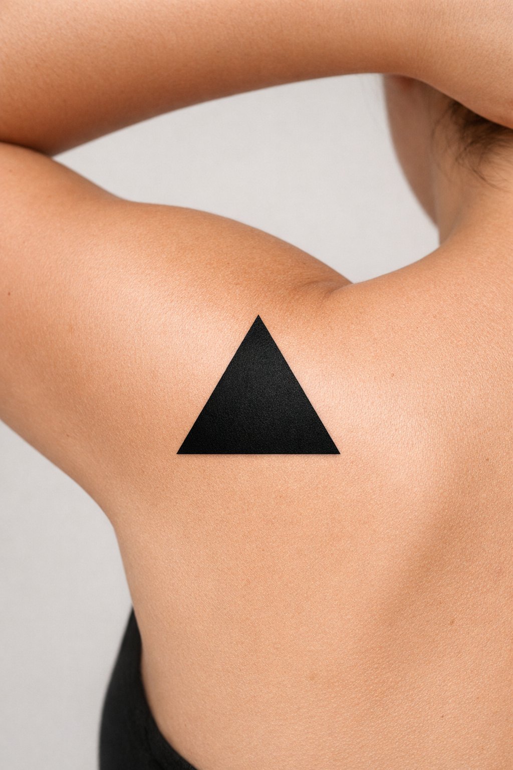 Close-up of a solid black geometric triangle tattoo on the shoulder blade. The arm is slightly raised behind the head, tightening the skin to show the design's crisp edges against a plain wall.