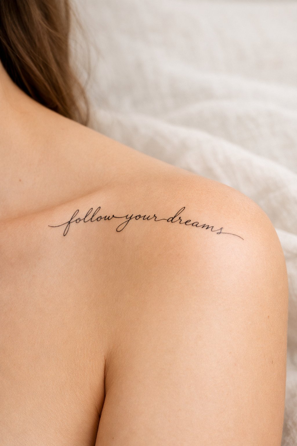 Detail shot of a cursive script tattoo following the natural curve of the shoulder. The text is thin and elegant, resting on bare skin with a linen sheet visible in the blurred background.