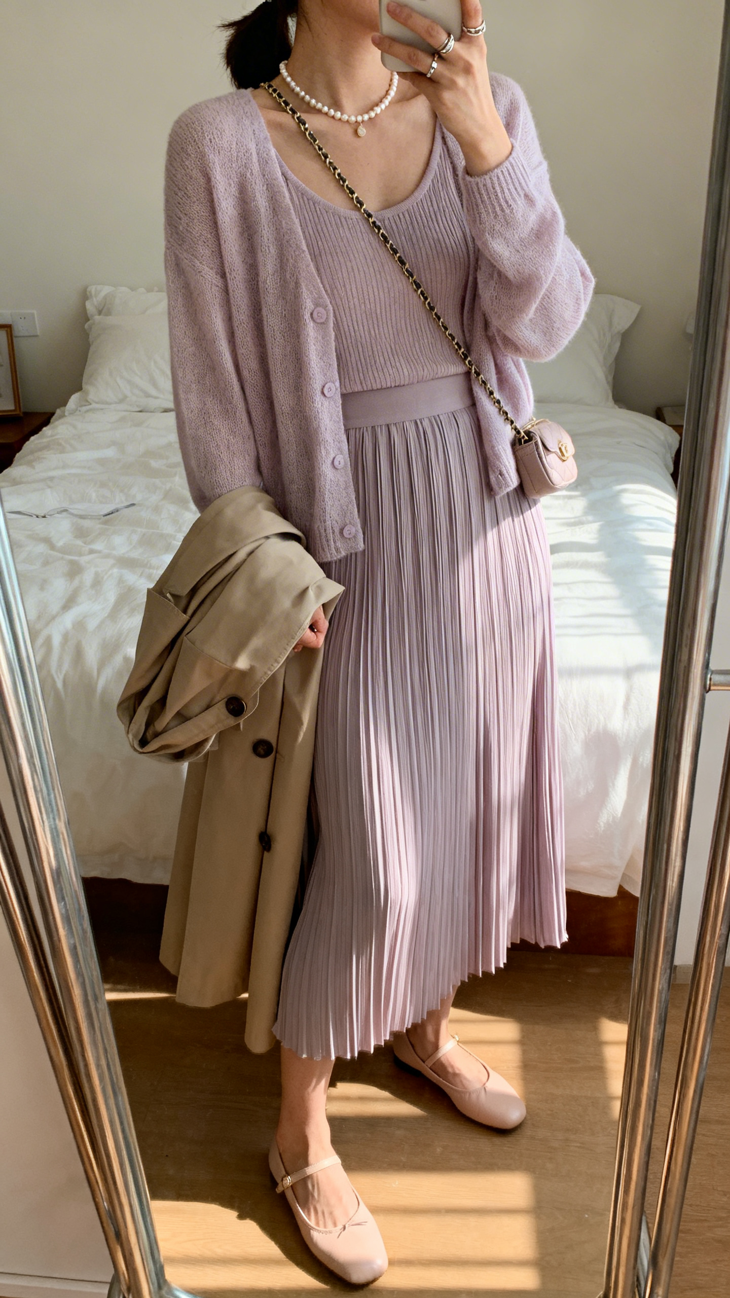 Bedroom mirror selfie with morning light: a woman in a fine-knit cardigan set (matching tank and cardi) in pastel, micro-pleated midi skirt in a complementary shade, ballet flats, small chain-strap crossbody, pearl necklace and dainty rings, trench over arm optional, side angle, face not visible, iPhone photo quality.