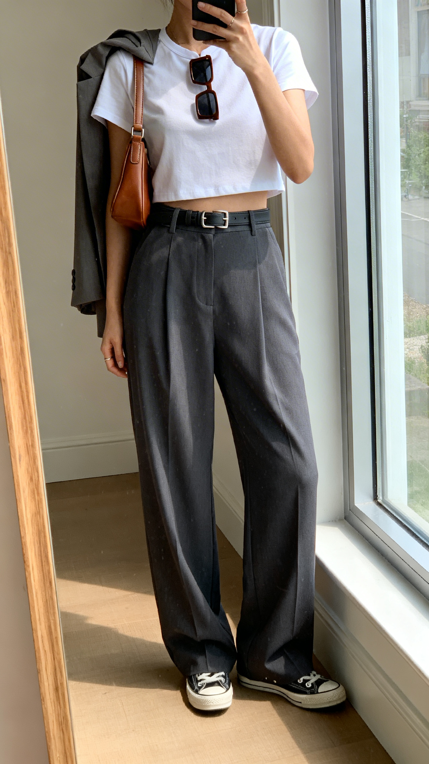 Full-length mirror selfie by window: a woman in high-waisted wide-leg trousers in charcoal with a white cropped crewneck tee, retro sneakers, subtle-hardware belt, rectangular sunglasses, leather shoulder bag, optional lightweight blazer carried, cropped at shoulders, face not visible, iPhone photo quality.