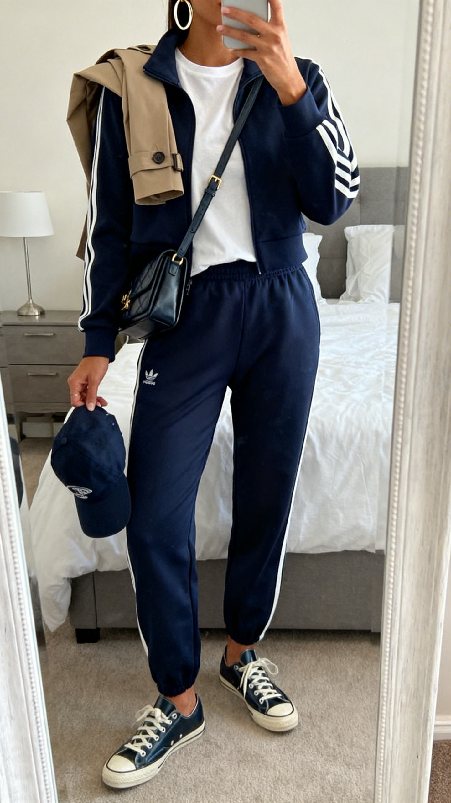 Full-length mirror selfie in bedroom: a woman in a coordinating navy track jacket and pants, high-quality white tee, leather low-top sneakers, structured crossbody, baseball cap in hand, oversized hoops, optional trench draped over shoulders, cropped at shoulders, face not visible, iPhone photo quality.