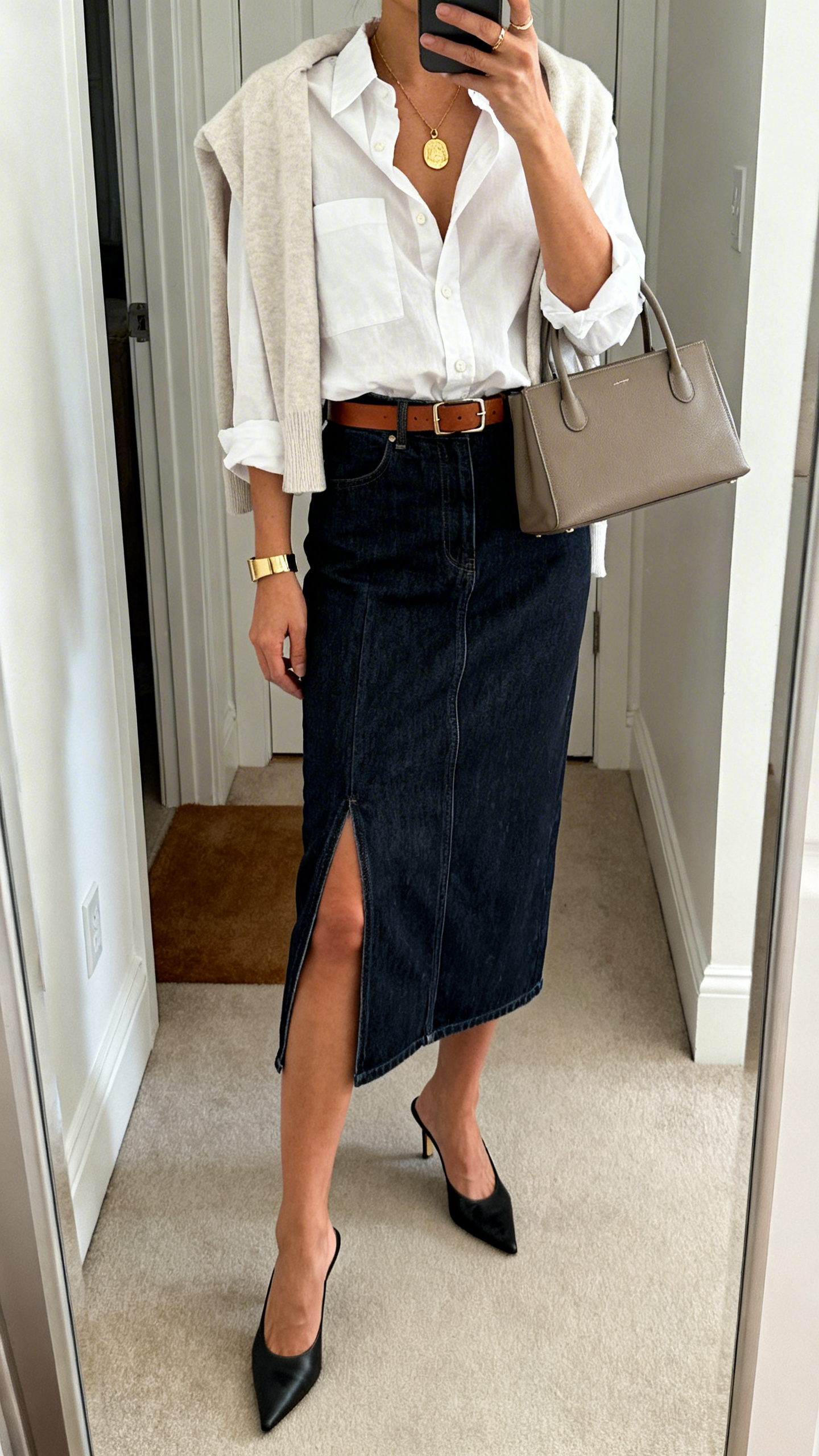Mirror selfie in entryway: a woman wearing a dark-wash denim midi skirt with front slit, relaxed men’s-inspired white button-up half-tucked, kitten-heel mules, slim leather belt, top-handle bag, gold pendant necklace and cuff bracelet, light cardigan draped over shoulders optional, cropped at neck, face not visible, iPhone photo quality.