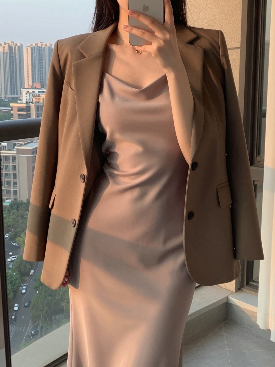 iPhone mirror selfie of a woman in a slip dress with a blazer over it, holding her phone low while holding the blazer's lapel, cropped at NECK so face doesn't show, in a city apartment, evening golden hour, iPhone photo quality.