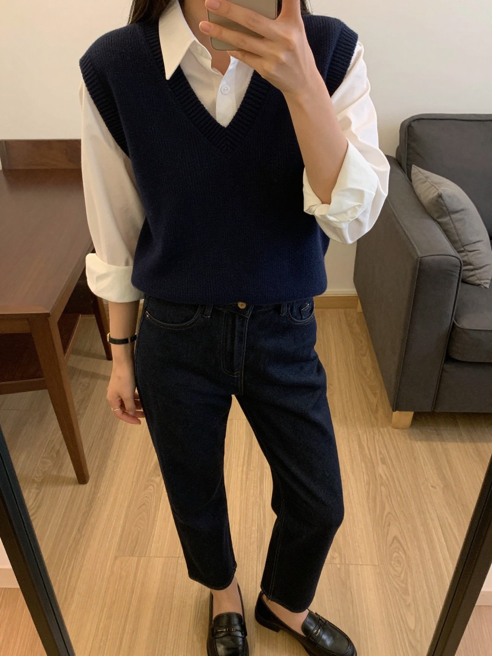 iPhone mirror selfie of a woman in a navy knit vest over a white button-down with dark straight jeans and loafers, adjusting her collar, cropped at NECK so face doesn't show, in a city apartment, warm moody lighting, iPhone photo quality.
