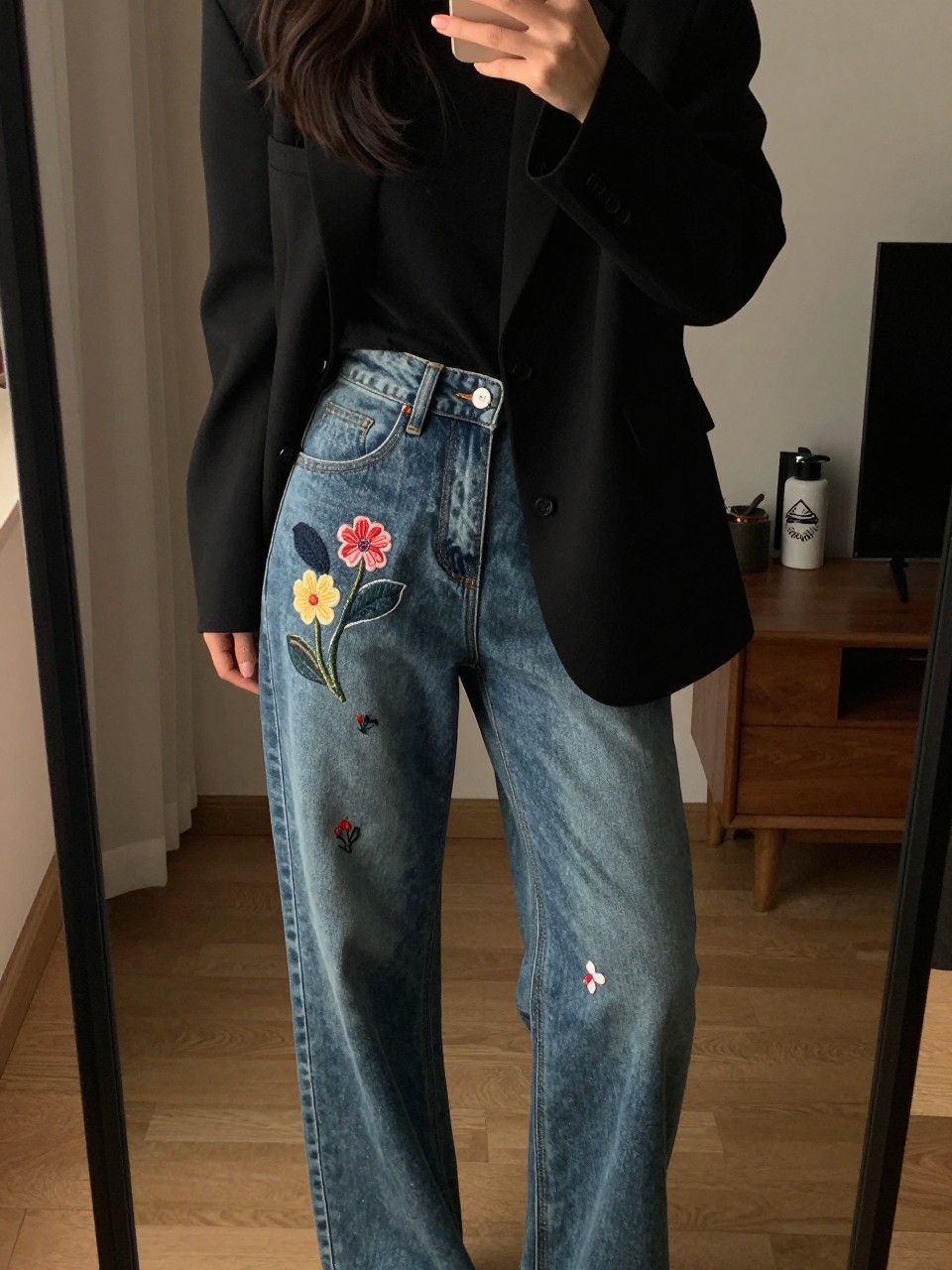iPhone mirror selfie of a woman in embroidered jeans and a blazer, showcasing the pant detail, cropped at NECK so face doesn't show, in a city apartment, warm moody lighting, iPhone photo quality.