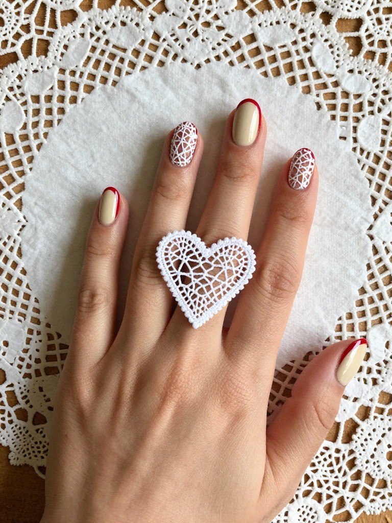 Top-down view of hands resting on an antique lace doily. The nails are an oval shape with a creamy, retro red polish. A fine, white lace pattern is painted over all nails. On the ring finger, the lace forms a distinct, intricate heart shape in the center. Soft, diffused light highlights the delicate details. The mood is vintage and romantic.