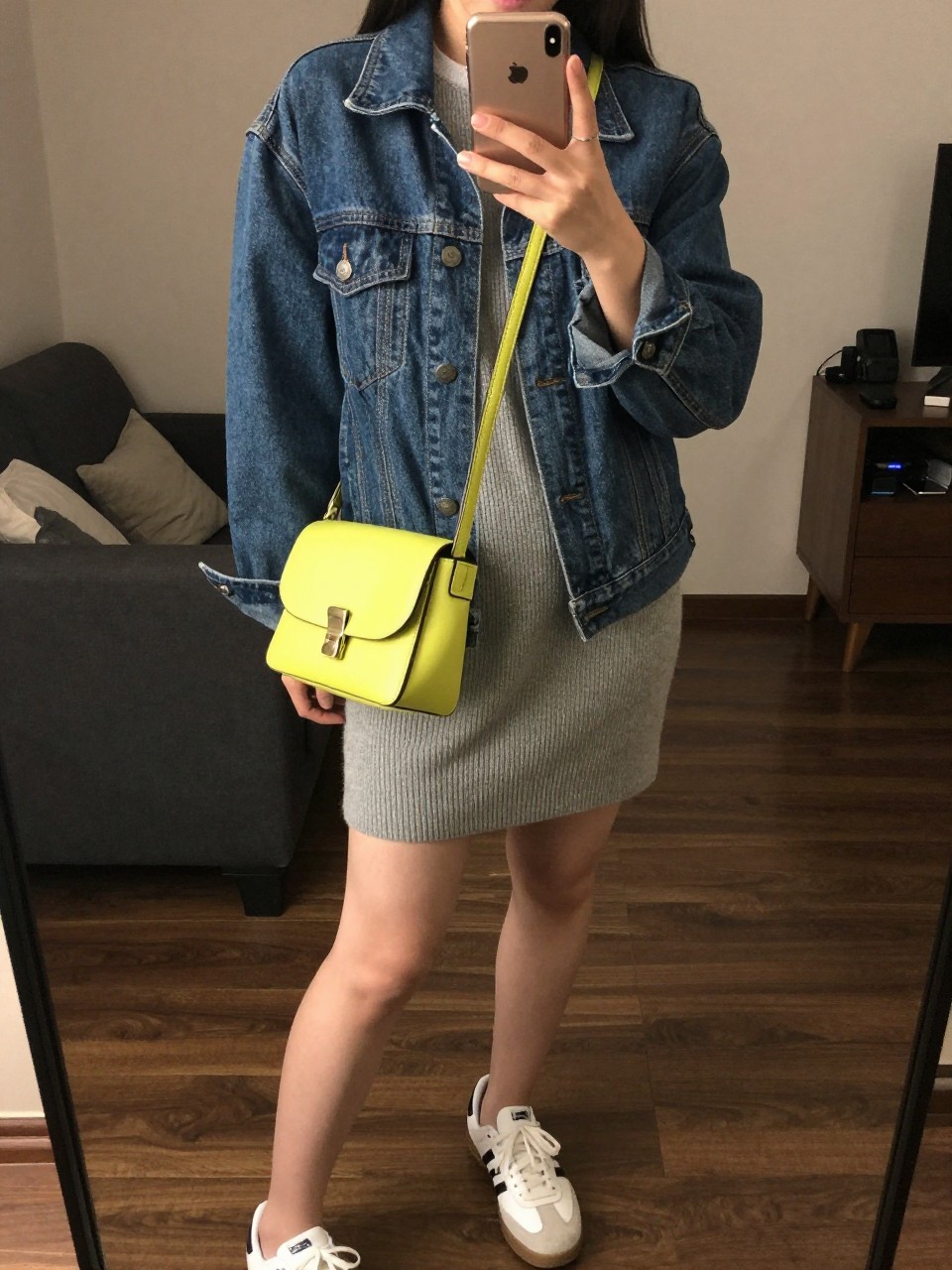 iPhone mirror selfie of a woman wearing the denim jacket over a grey sweater dress with white sneakers and a bright crossbody bag, posed looking down at her phone, cropped at NECK so face doesn't show, in a city apartment, warm moody lighting, iPhone photo quality.