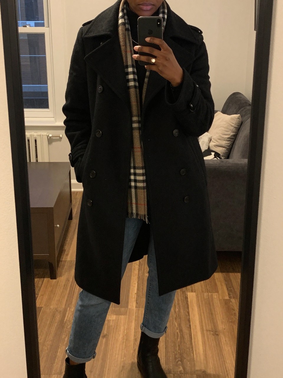 iPhone mirror selfie of a Black woman wearing a black peacoat with a plaid scarf and blue jeans, standing casually with her phone at her side, cropped at NECK so face doesn't show, in a city apartment, warm moody lighting, iPhone photo quality.