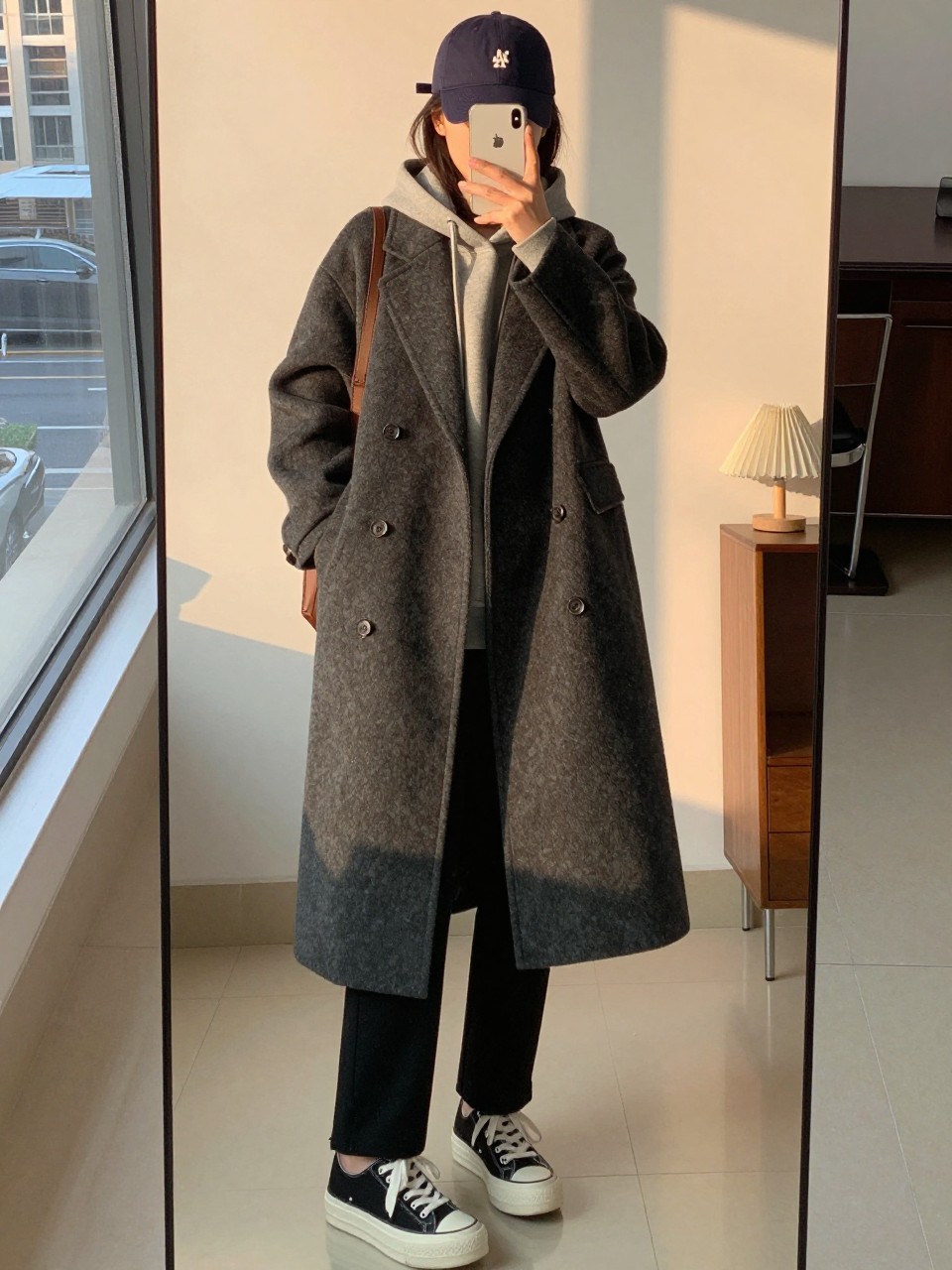 iPhone mirror selfie of a woman in a long charcoal wool coat over a gray hoodie with black trousers and platform sneakers, wearing a baseball cap, cropped at NECK so face doesn't show, in a city apartment, evening golden hour, iPhone photo quality.