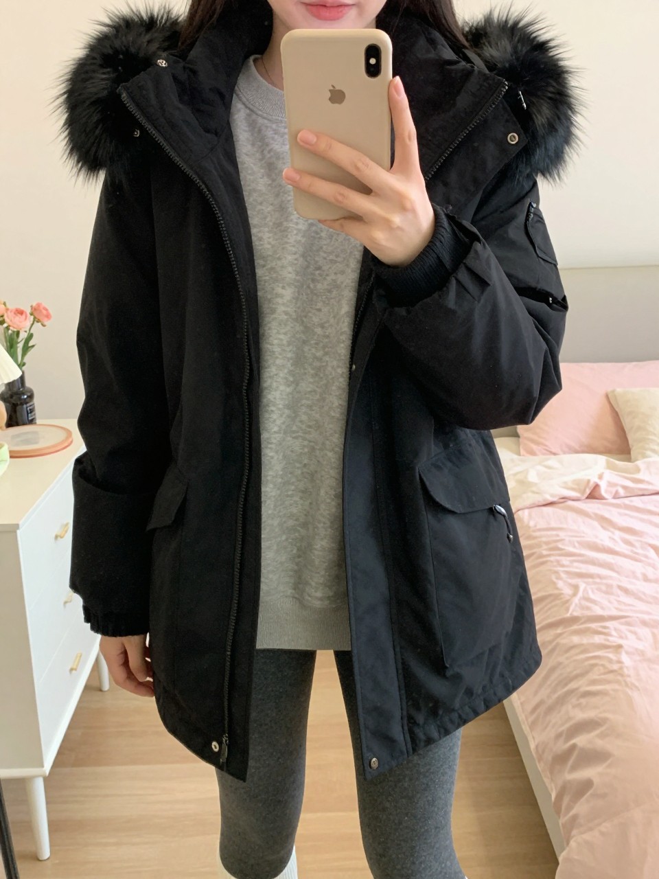 iPhone mirror selfie of a woman wearing a black parka with faux fur trim over a gray sweatshirt and leggings, standing with her phone in front, cropped at NECK so face doesn't show, in a bright feminine-themed bedroom, warm moody lighting, iPhone photo quality.