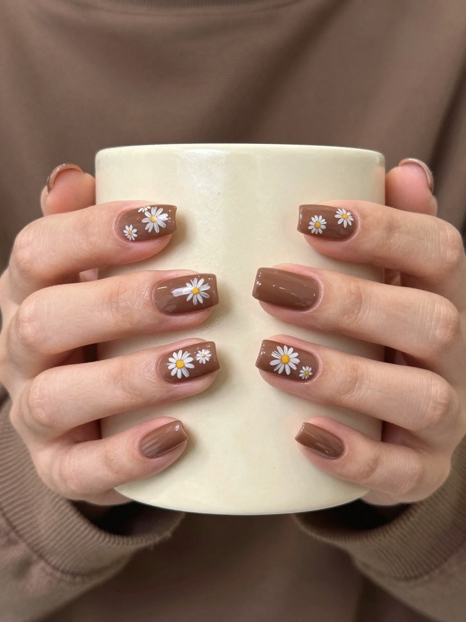 Overhead, top-down view of feminine hands with almond nails resting on a textured, cream-colored ceramic mug. The nails are a smooth milk chocolate brown. Delicate white line-art daisies are scattered across each nail. The lighting is warm and cozy, like in a café.