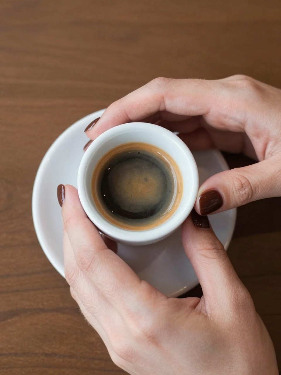 Bird's-eye view of hands wrapped around a small, white ceramic espresso cup. The nails are a short, rounded shape in a high-gloss, dark chocolate brown. The café setting is warm and intimate.