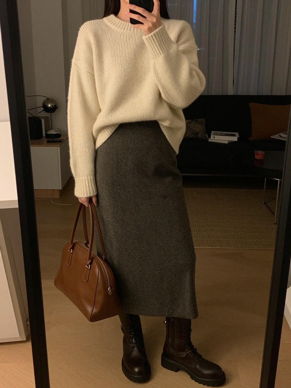 iPhone mirror selfie of a woman wearing the chunky cream sweater partially tucked into a herringbone midi skirt with lug-sole boots and a brown leather bag, posed with her hand adjusting her sweater, cropped at NECK so face doesn't show, in a city apartment, warm moody lighting, iPhone photo quality.