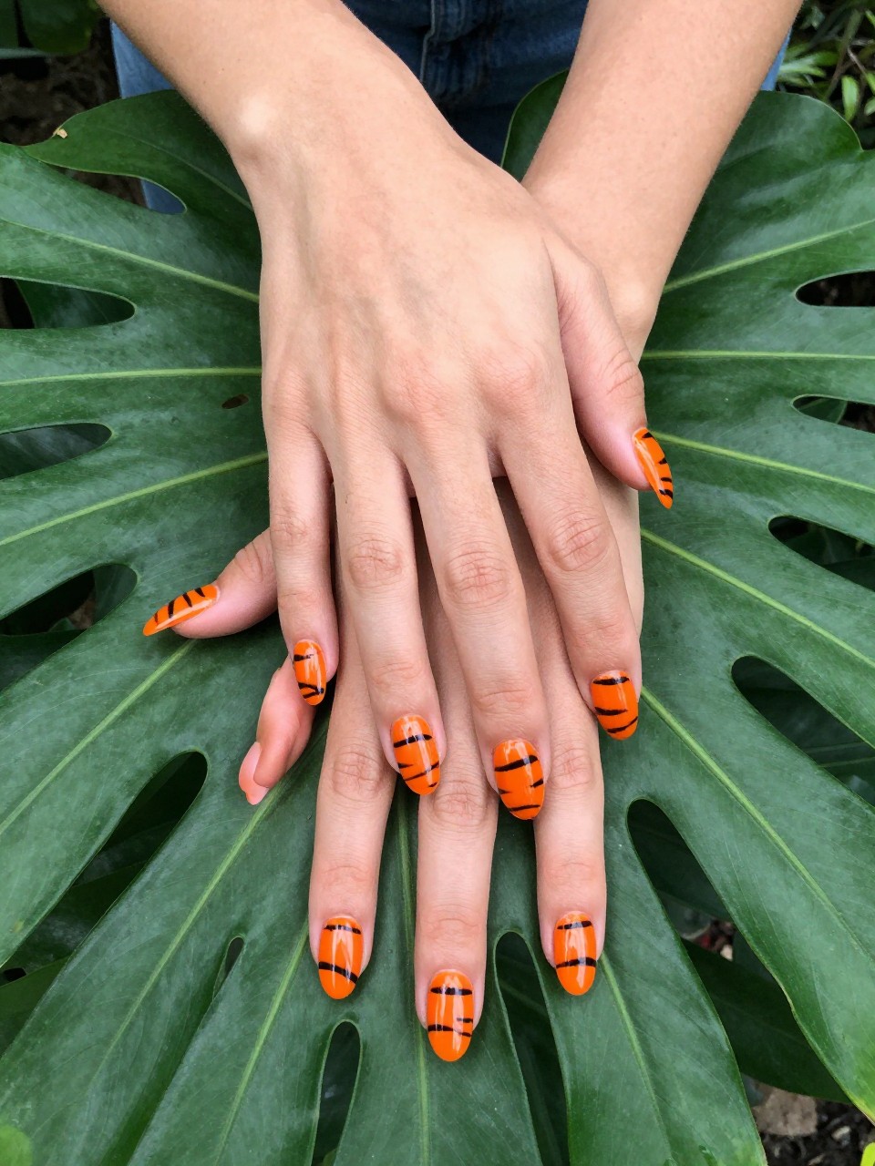 Low-angle view of hands resting on a lush green tropical leaf. The nails are glossy orange with sharp, black tiger stripes. The look is wild and powerful.