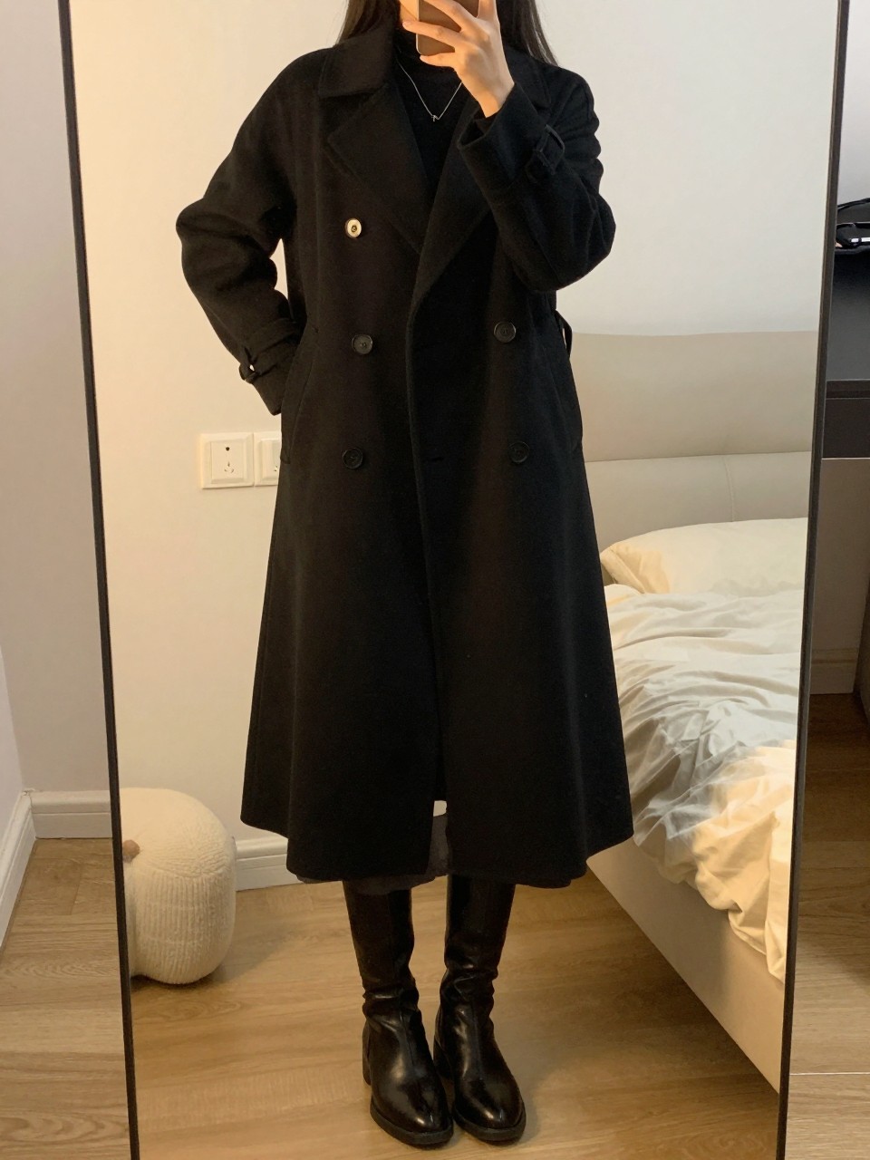 iPhone mirror selfie of a woman wearing a double-breasted black coat over a midi skirt and knee-high boots, standing with one hand on her hip, cropped at NECK so face doesn't show, in a feminine-themed bedroom, evening golden hour light, iPhone photo quality.