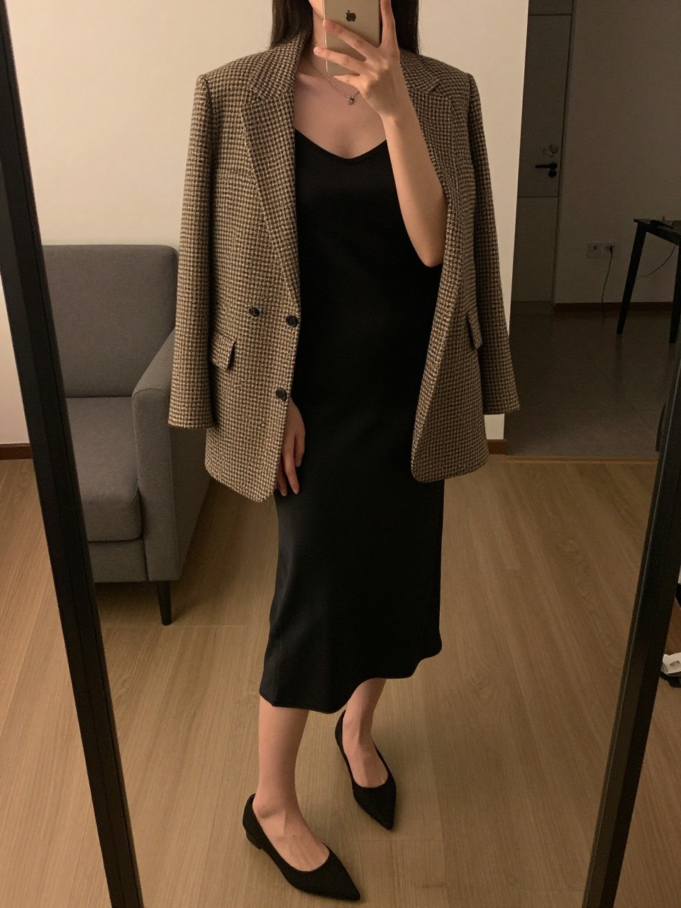 iPhone mirror selfie of a woman in a houndstooth blazer over a black camisole and matching slip skirt with pointed flats, standing with phone near her shoulder, cropped at NECK so face doesn't show, in a city apartment, warm moody lighting, iPhone photo quality.