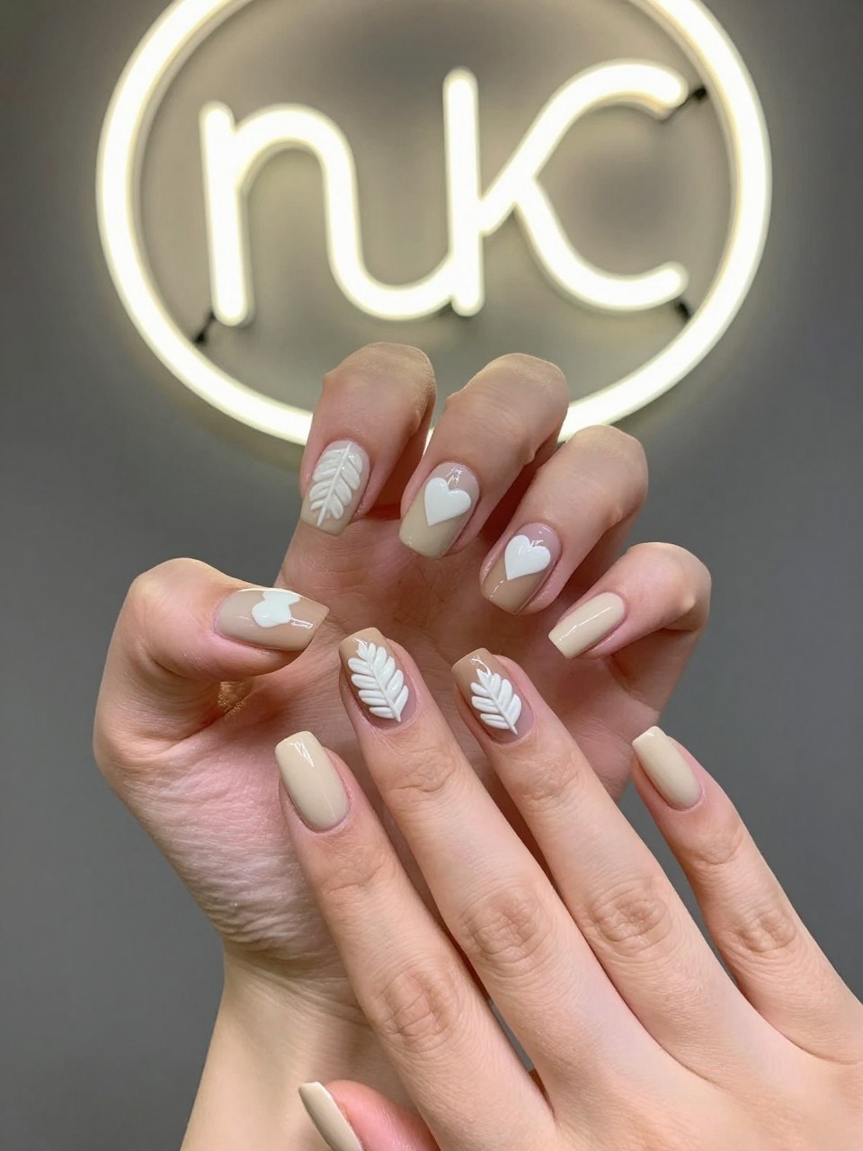 Top-down view of hands next to a neon studio light. The nails are a creamy light brown. Each nail has a textured white gel leaf or heart design, mimicking latte art. The vibe is cozy and whimsical.