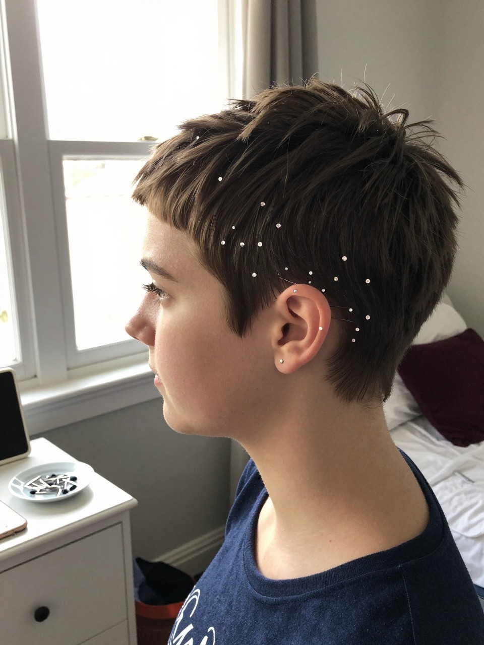 Photo of a teenage white girl wearing an accessorized pixie with sparkly pins, side profile view showing the pins clustered near the ear, setting: a bedroom with natural light from a window, lighting: bright, soft window light, containing a small dish with spare pins on a dresser, iPhone photo quality.