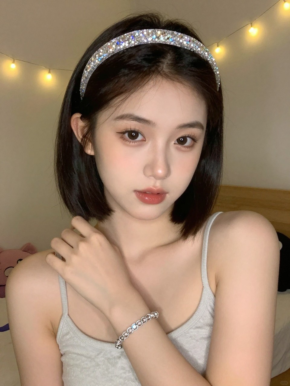 Photo of a teenage girl wearing a bob with a sparkly headband, front-facing view showing the headband, setting: a bedroom with fairy lights in the background, lighting: warm, soft artificial light, containing a matching bracelet on her wrist, iPhone photo quality.