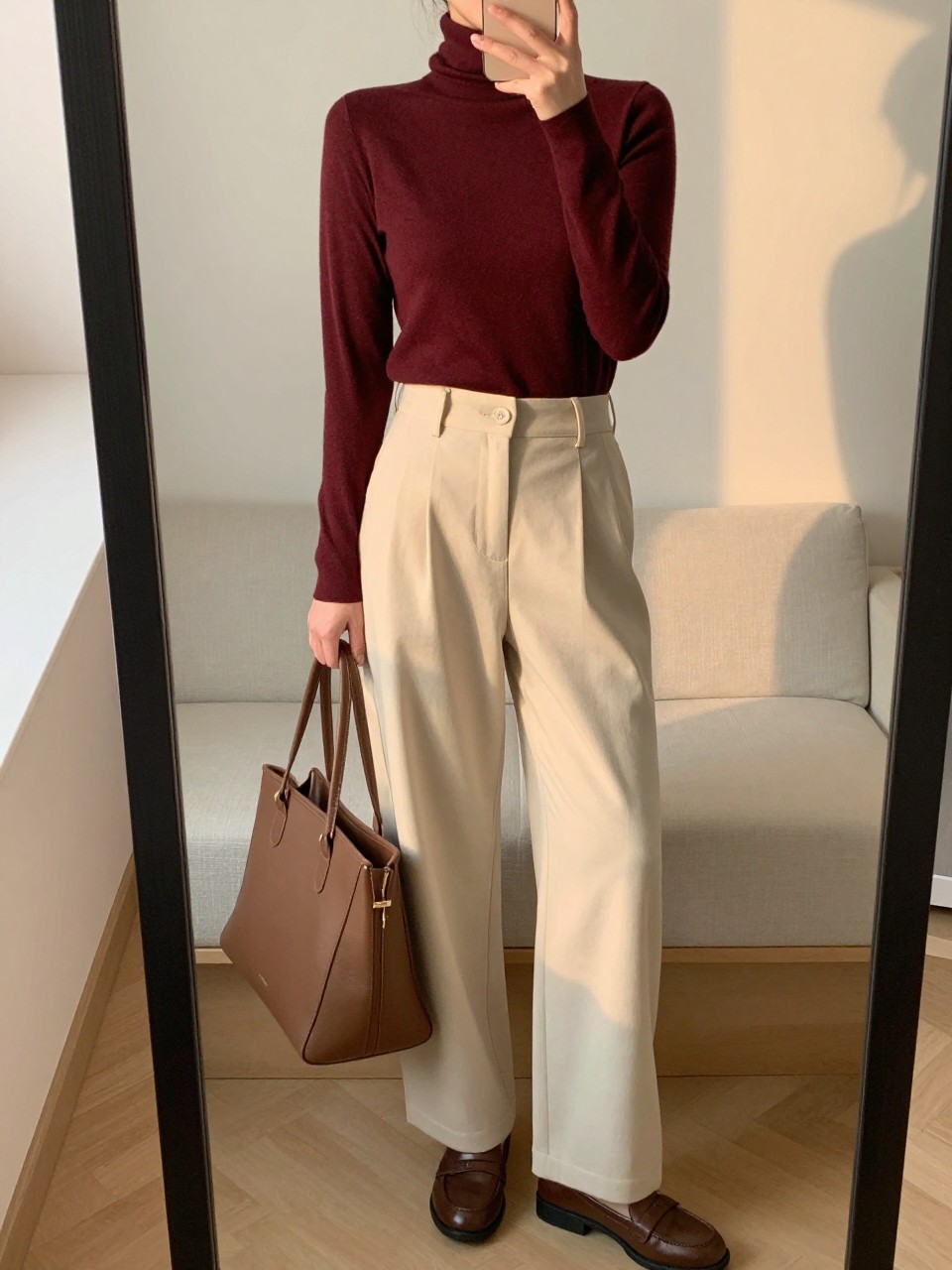 iPhone mirror selfie of a woman wearing the burgundy turtleneck tucked into cream wide-leg trousers with loafers and a large tote, posed looking slightly away from the mirror, cropped at NECK so face doesn't show, in a feminine-themed bedroom, evening golden hour, iPhone photo quality.