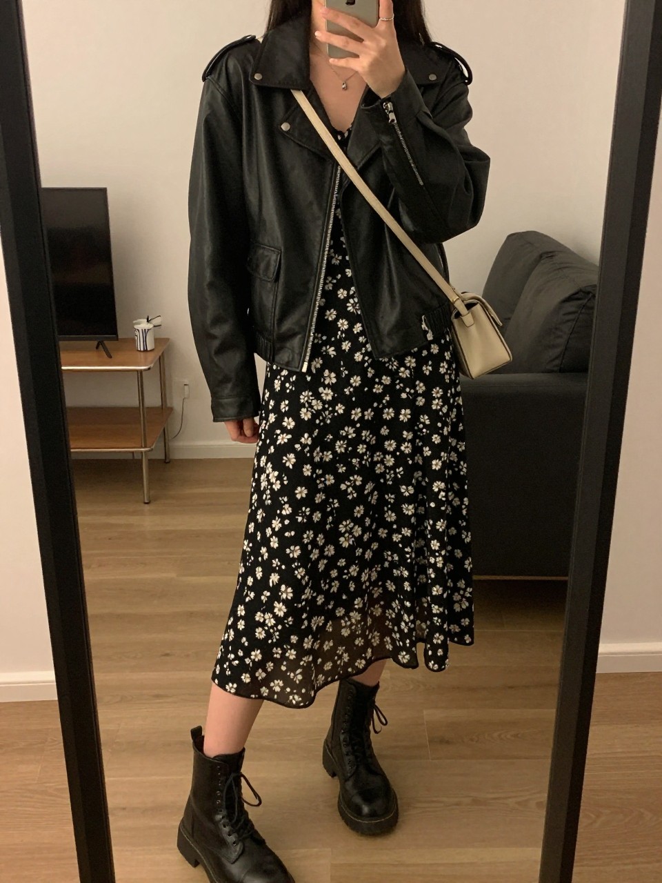 iPhone mirror selfie of a woman wearing the black leather jacket over a floral midi dress with combat boots and a crossbody bag, posed with one hand in her jacket pocket, cropped at NECK so face doesn't show, in a city apartment, warm moody lighting, iPhone photo quality.