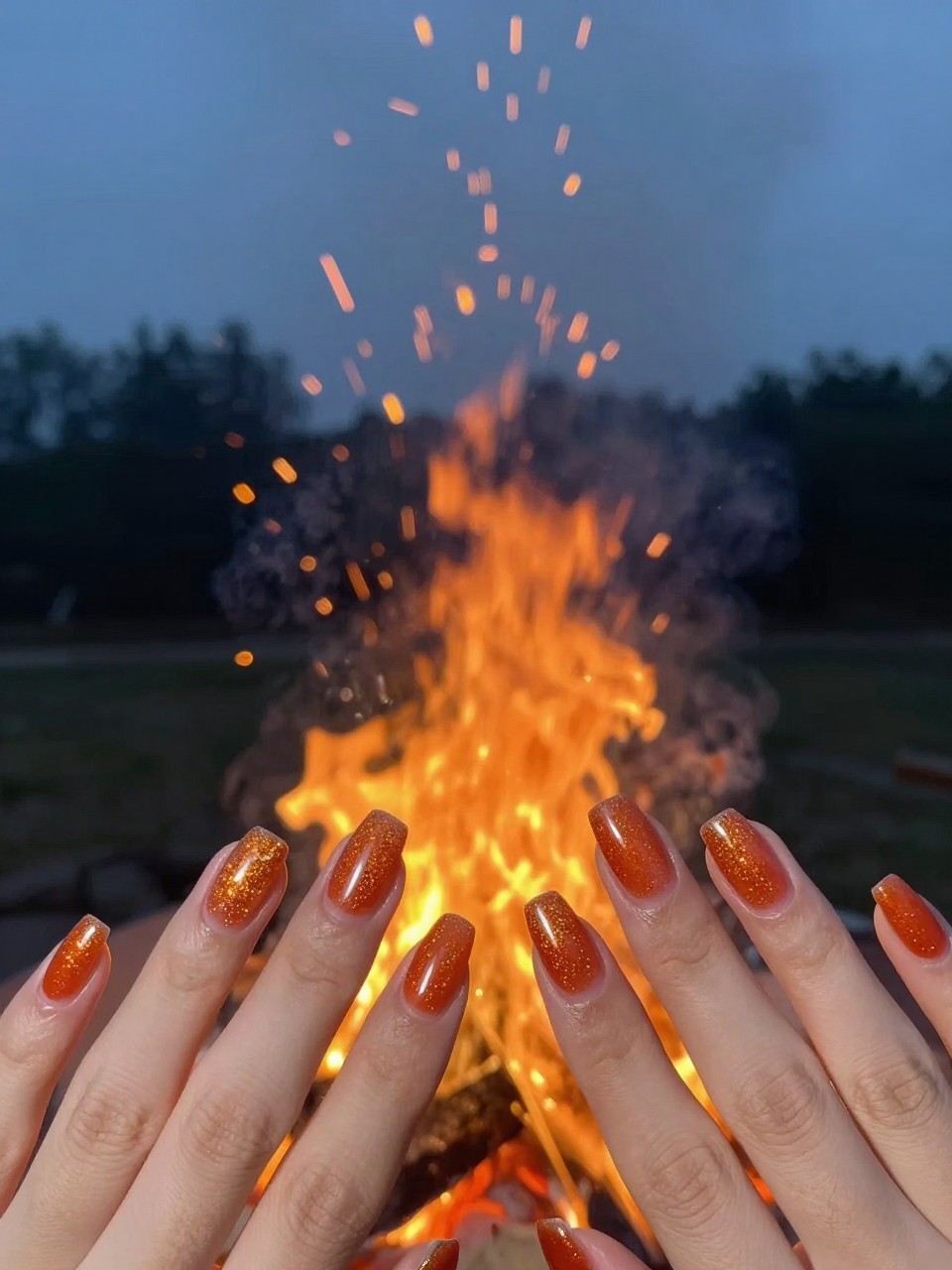 Image Prompt: Shot of hands at a bonfire at dusk, sparks flying upward. The natural nails are glossy with a concentrated orange glitter fade at the tips. The glitter mimics the rising embers.