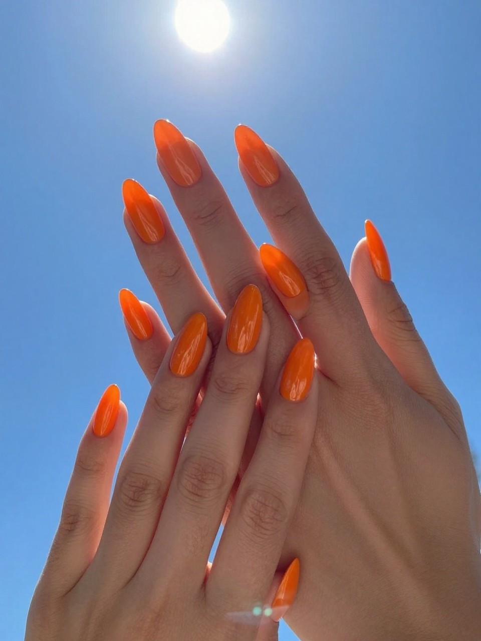 Overhead view of hands against a clear blue sky, sun directly overhead. The almond nails are a high-gloss, pure orange, so vibrant they seem to glow from within. The contrast with the blue sky is stunning.