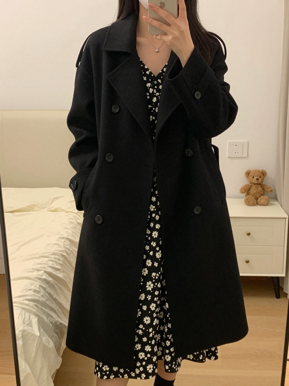 iPhone mirror selfie of a woman wearing a black swing coat over a printed dress, standing with one hand holding her phone at her side, cropped at NECK so face doesn't show, in a feminine-themed bedroom, warm moody lighting, iPhone photo quality.