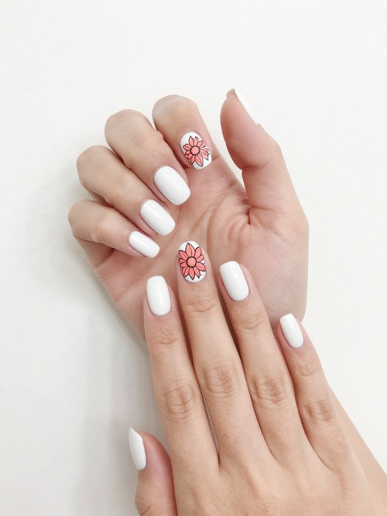 Hands on a pure white minimalist backdrop. The gel nails are glossy white. A single, graphic black-outline dahlia filled with coral color sits centered on each nail. The design is bold, artistic, and clean.