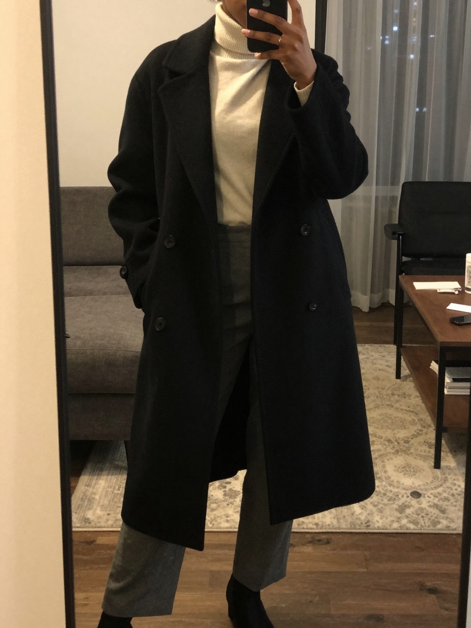 iPhone mirror selfie of a Black woman wearing a long black wool coat over a cream turtleneck and charcoal trousers, standing with her weight on one leg and hands casually in her coat pockets, cropped at NECK so face doesn't show, in a city apartment, warm moody lighting, iPhone photo quality.