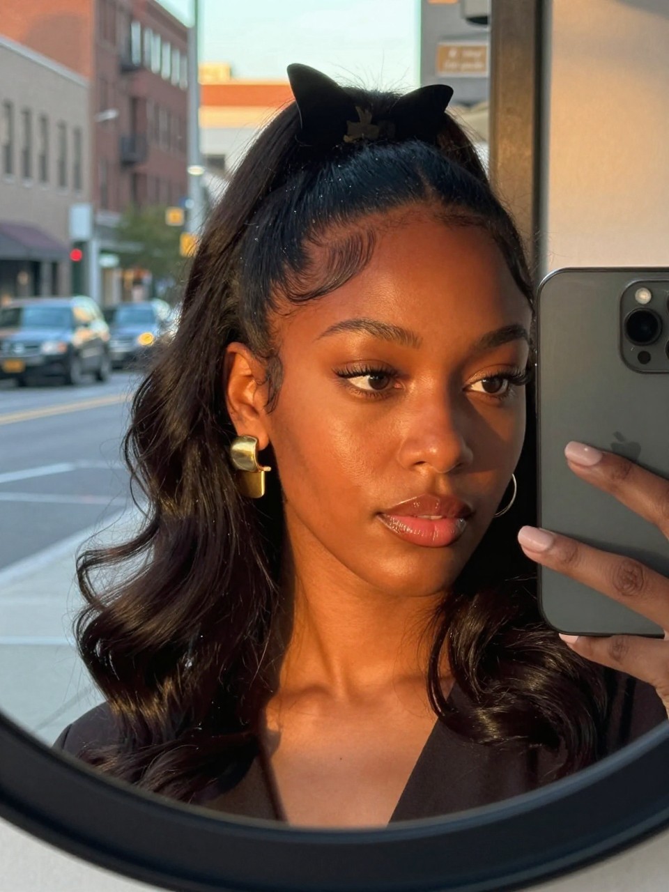 Photo of a black woman wearing a half-up butterfly clip style, candid mirror selfie with her phone visible, blurred city street background through a window, golden hour light warming her face, containing statement gold earrings peeking through her waves, iPhone photo quality.