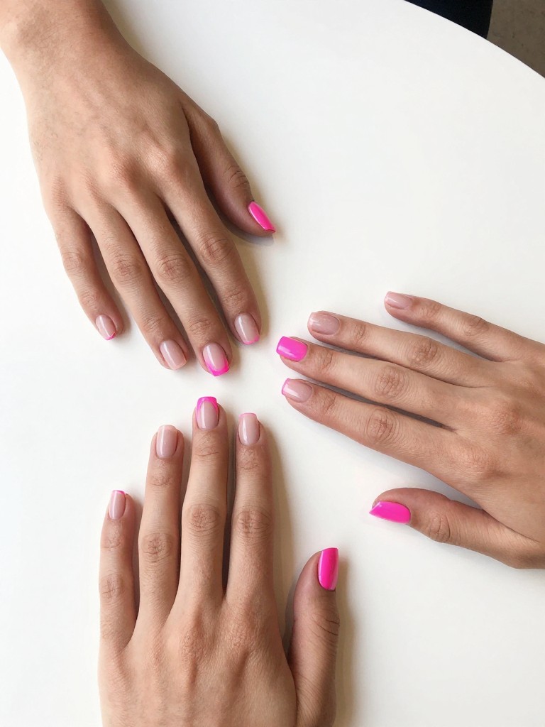 Top-down view of hands on a minimalist, white cafe table. The short, square nails are precisely split diagonally: half sheer nude, half matte neon pink. The contrast is striking in clear daylight.