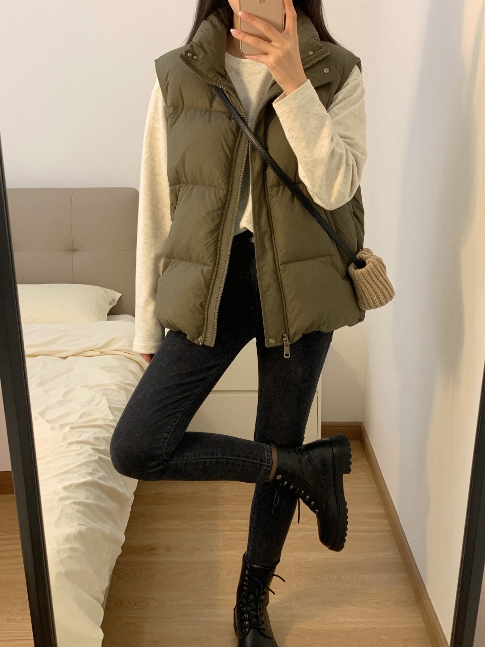 iPhone mirror selfie of a woman wearing the olive puffer vest over a cream long-sleeve tee with dark jeans and combat boots, holding a beanie and small crossbody, posed with her weight on one leg, cropped at NECK so face doesn't show, in a feminine-themed bedroom, evening golden hour, iPhone photo quality.