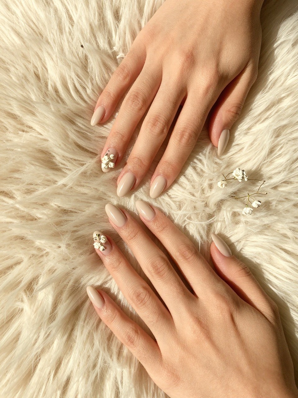 Overhead, top-down view of feminine hands with almond nails resting on a textured, cream-colored fur rug. The nails are a smooth Sheer Beige. Delicate with Pressed Baby’s Breath scattered across each nail. The lighting is warm and cozy, like in a nail salon.