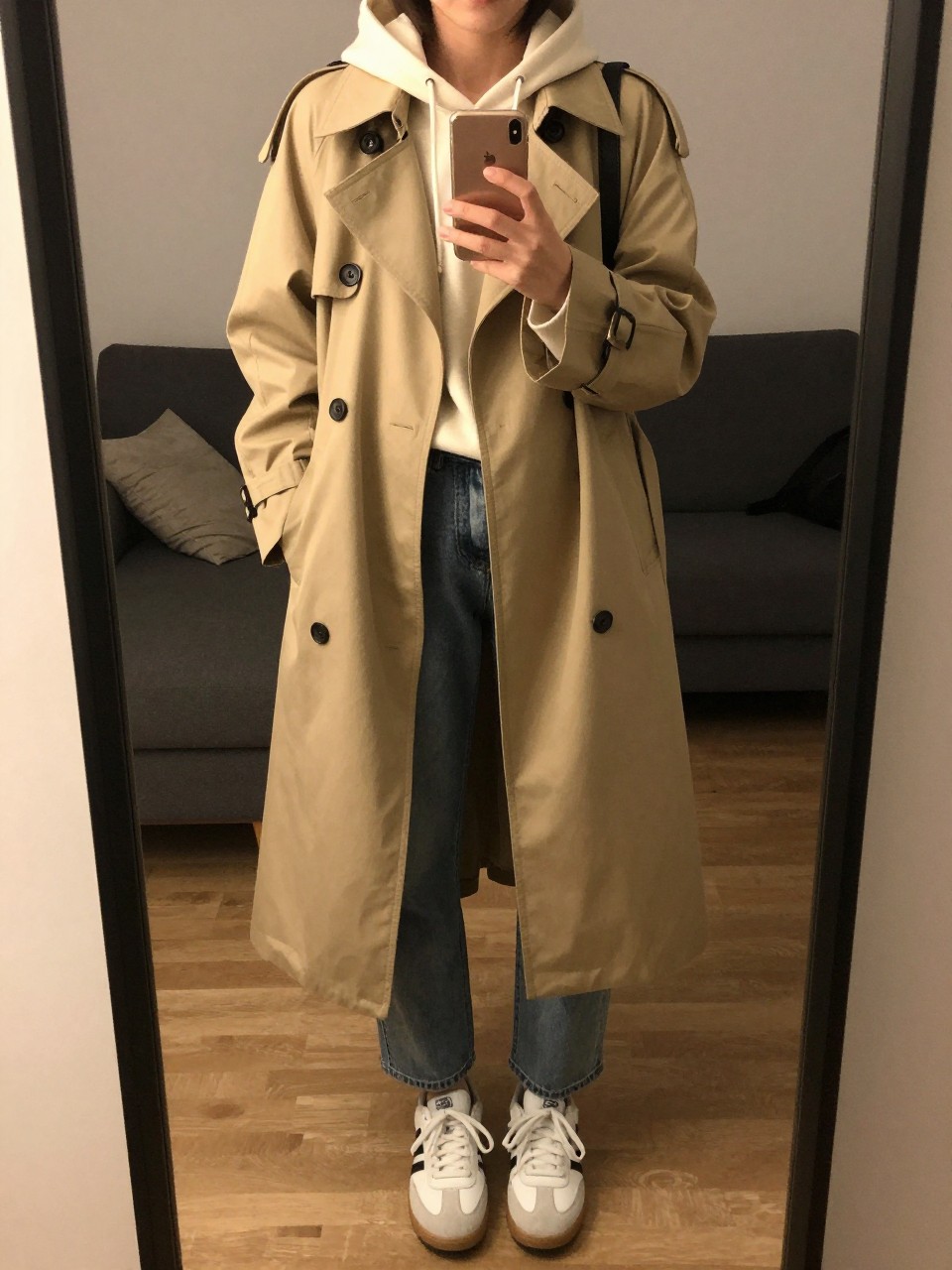 iPhone mirror selfie of a woman in a beige trench coat over a cream hoodie and medium-wash jeans with white sneakers, standing with phone at chest level, cropped at NECK so face doesn't show, in a city apartment, warm moody lighting, iPhone photo quality.
