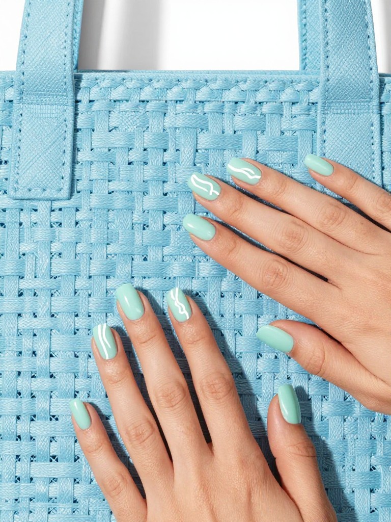 Top-down view of hands resting on a light blue, woven beach bag. The nails are a short, square shape in a soft mint green. A single, hand-painted wavy white line crosses each nail about halfway down. The lighting is bright and clean, emphasizing the crisp design.