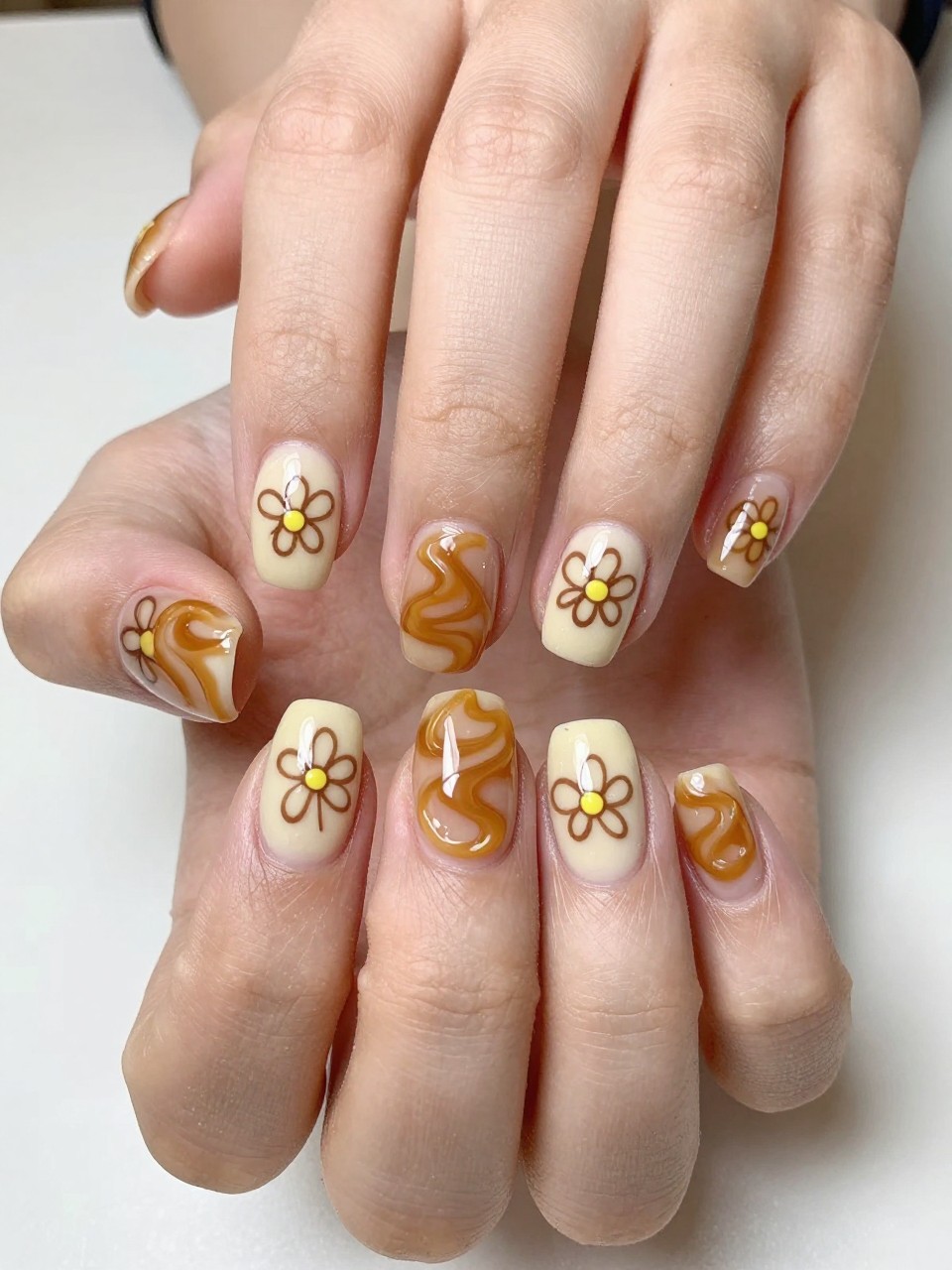 Top-down view of hands next to a caramel dessert. The nails have a swirled caramel and cream base. Simple brown-outline buttercups with tiny yellow centers are dotted across each nail. The look is sweet and cheerful.