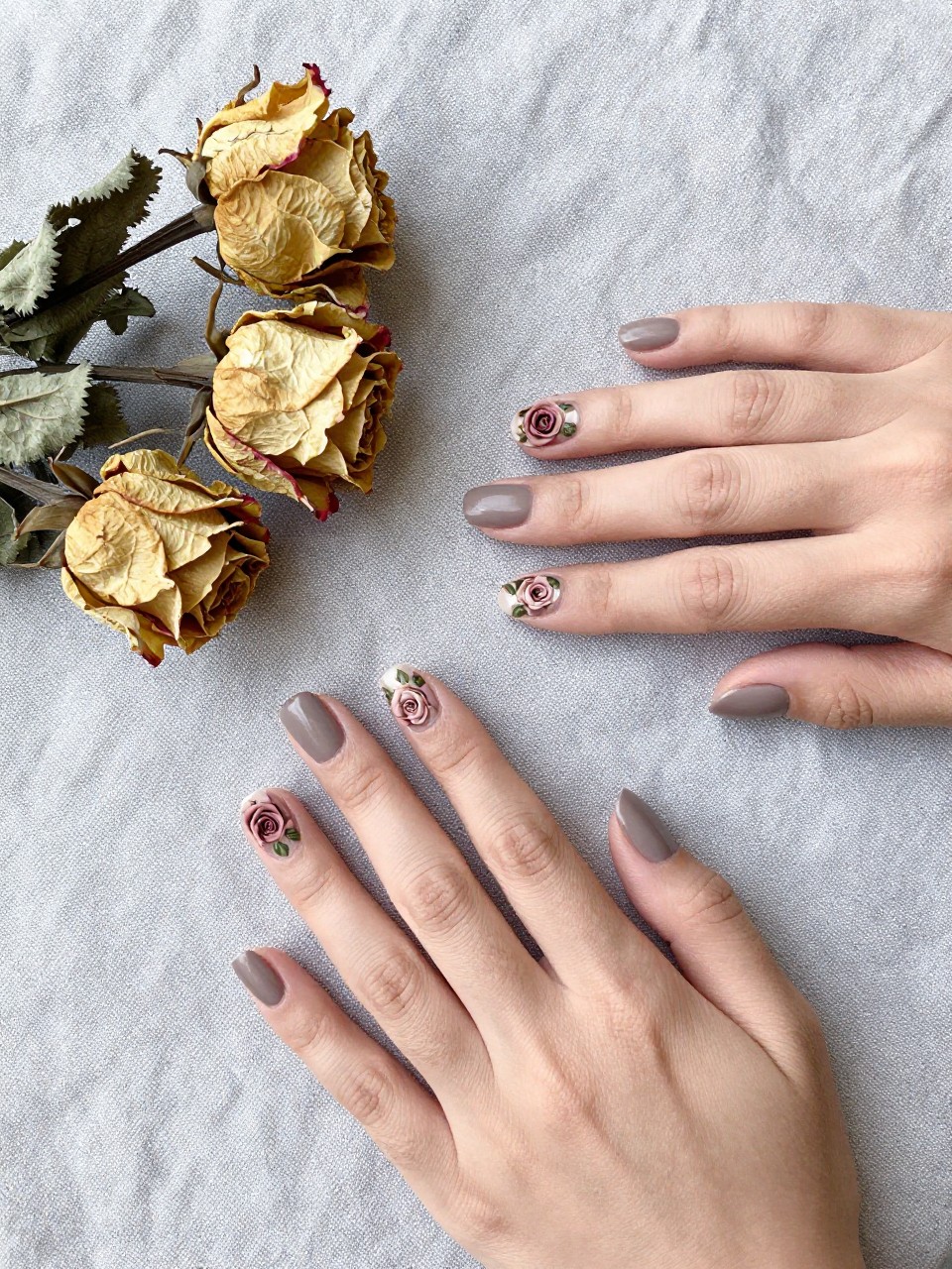 Bird's-eye view of hands on a grey linen tablecloth next to a dried rose bouquet. The squoval nails are a matte taupe. Each nail features a single, small dusty rose rosette with subtle green leaves. The mood is serene and artistic.