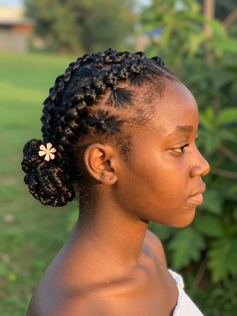 Photo of a teenage black girl wearing a braided accent on short hair, side profile view showing the small braid, setting: a garden with soft green background, lighting: warm golden hour light, containing a small flower tucked near the braid, iPhone photo quality.