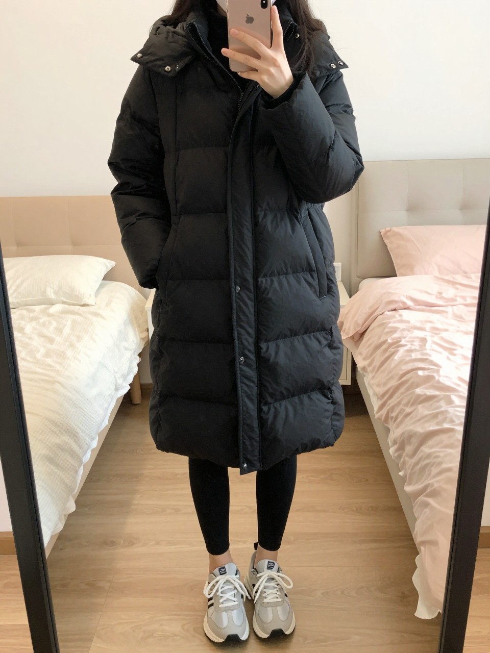 iPhone mirror selfie of a woman wearing a sleek black puffer coat with leggings and sneakers, standing with her weight shifted and phone low, cropped at NECK so face doesn't show, in a bright feminine-themed bedroom, warm moody lighting, iPhone photo quality.