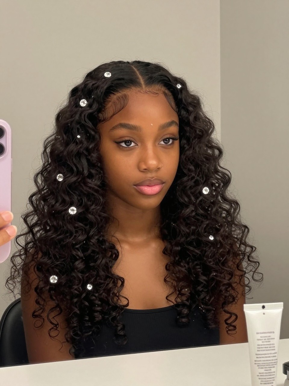 Photo of a black girl wearing defined curls with crystal pins, front view showing pins scattered through the hair, in soft indoor lighting against a plain background, containing a tube of curl cream visible on the counter, iPhone photo quality.
