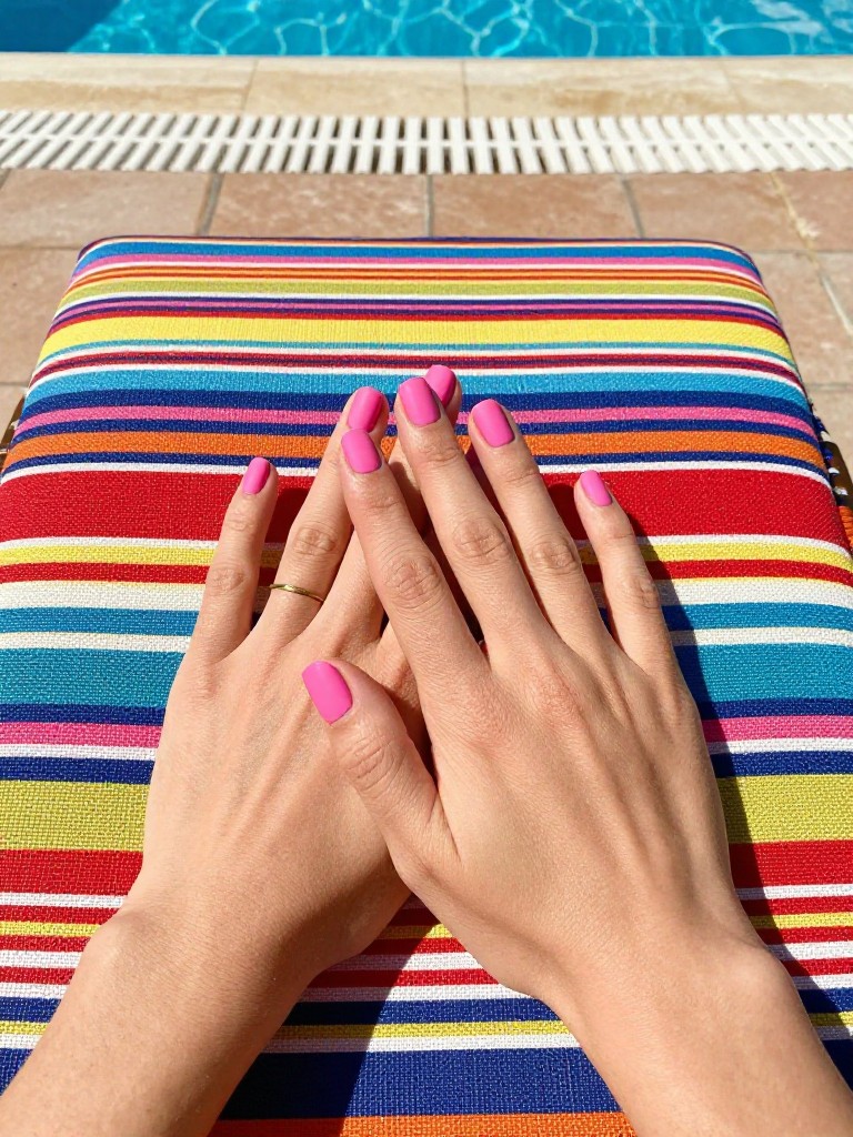 Bird's-eye view of hands resting on a colorful, patterned resort lounger. The nails are a short square shape in a flat, matte hot pink. The setting is a vibrant pool deck under the bright midday sun.