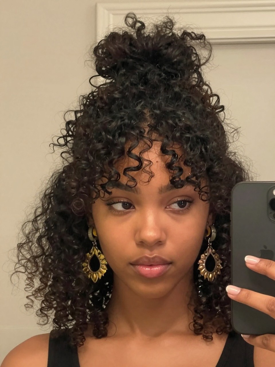 Photo of a black girl wearing a pineapple updo with curly bangs, front-facing candid mirror selfie showing the height and face-framing pieces, in soft indoor lighting, containing statement earrings peeking through the curls, iPhone photo quality.