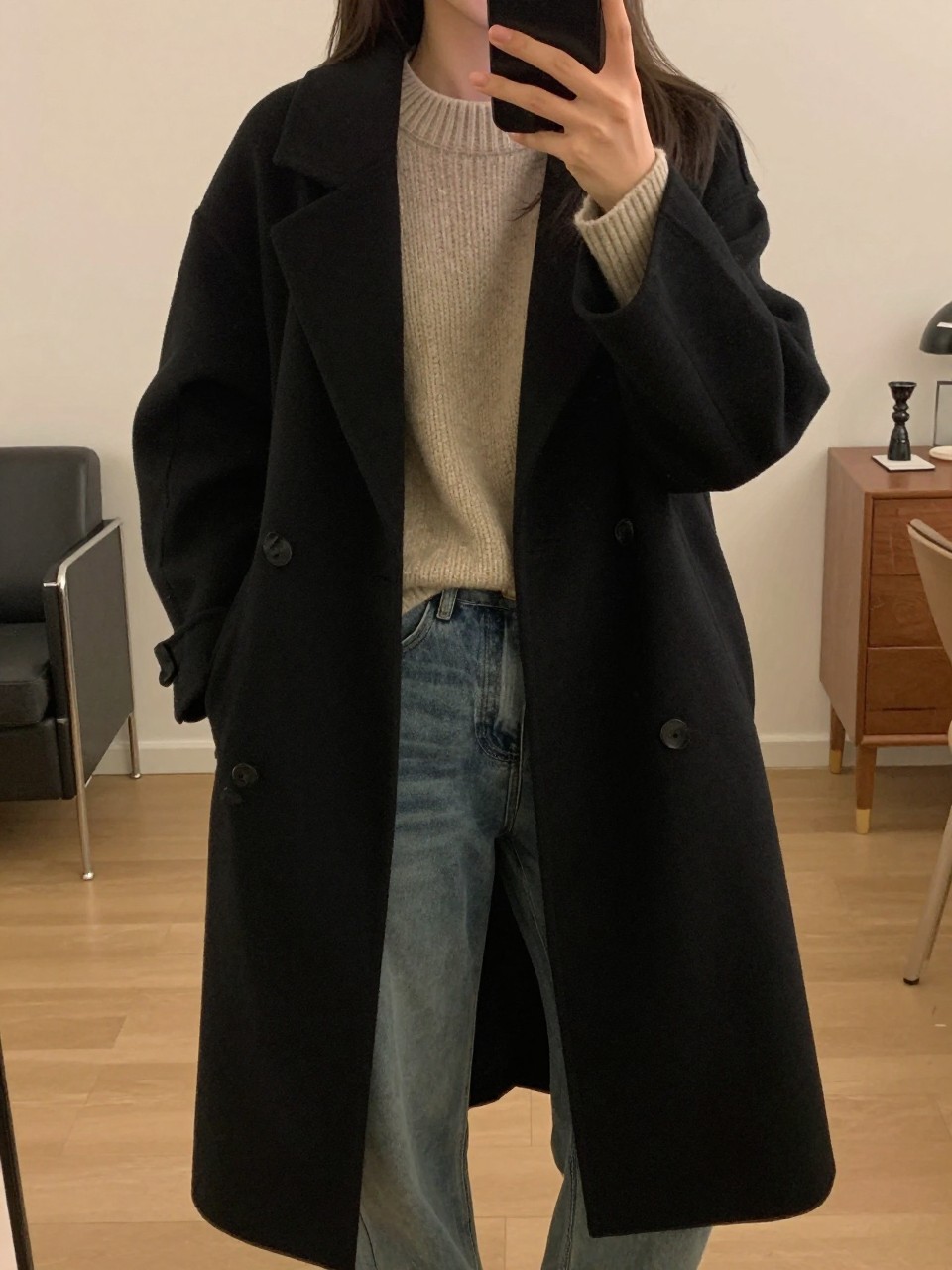iPhone mirror selfie of a woman wearing a black cocoon coat over a chunky oatmeal sweater and jeans, posed casually with her phone low, cropped at NECK so face doesn't show, in a city apartment, warm moody lighting, iPhone photo quality.