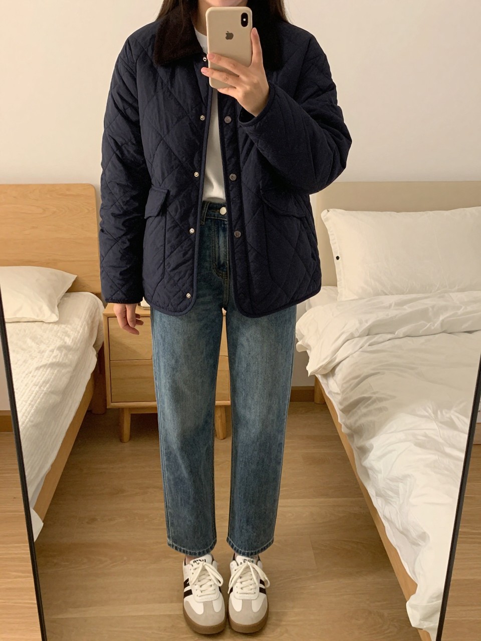 iPhone mirror selfie of a woman in a navy quilted jacket over a white tee and straight-leg jeans with white sneakers, standing with one foot forward, phone at chest level, cropped at NECK so face doesn't show, in a feminine-themed bedroom, evening golden hour, iPhone photo quality.